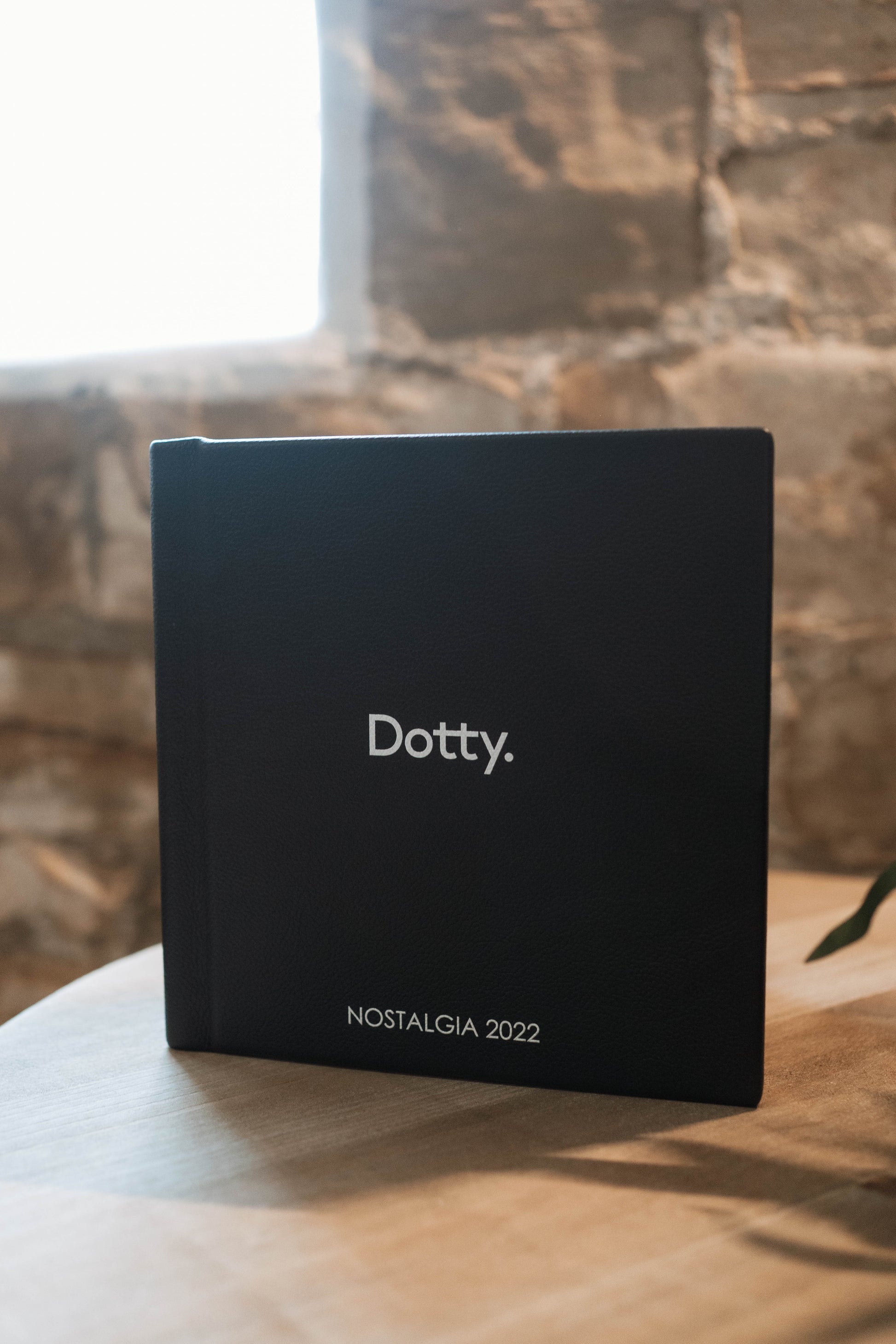 A black hardcover album standing upright on a wooden table, with the word 'Dotty.' printed in white near the center and 'NOSTALGIA 2022' near the bottom.