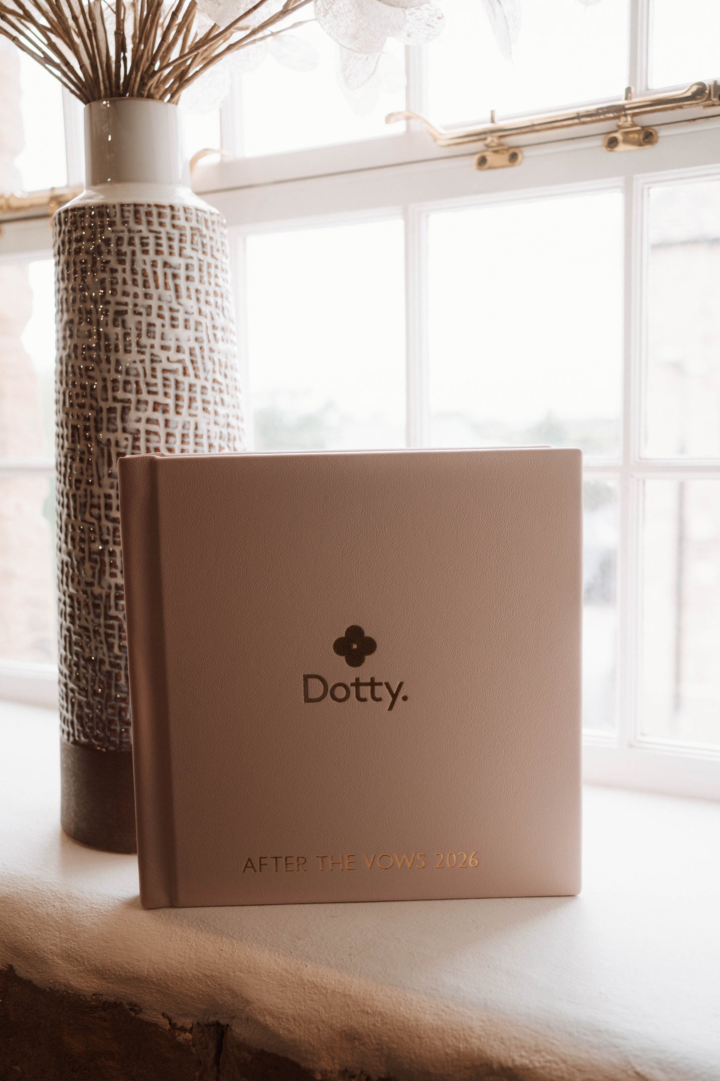 A square Dotty wedding album in a warm blush tone sits on a windowsill beside a tall textured vase with dried stems.