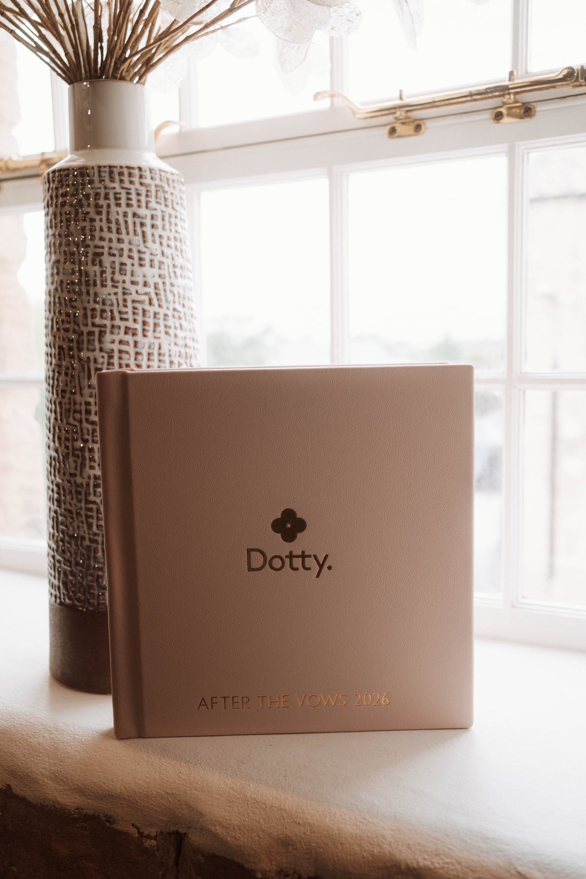 A square Dotty wedding album in a warm blush tone sits on a windowsill beside a tall textured vase with dried stems.