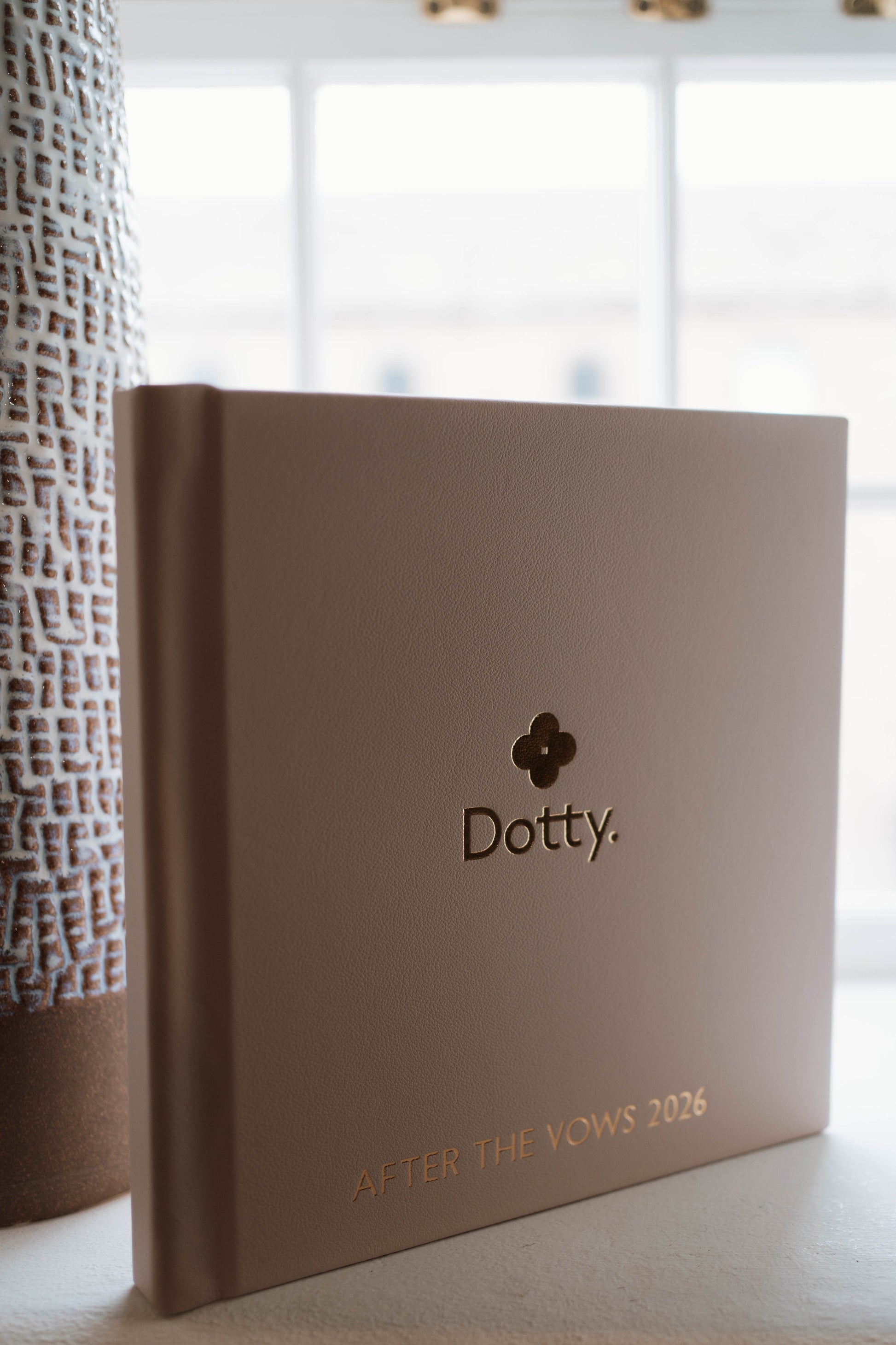 A square hardcover photo book titled 'Dotty. AFTER THE VOWS 2026' in a warm blush tone, standing on a light surface with a softly blurred bright window and textured vase in the background.