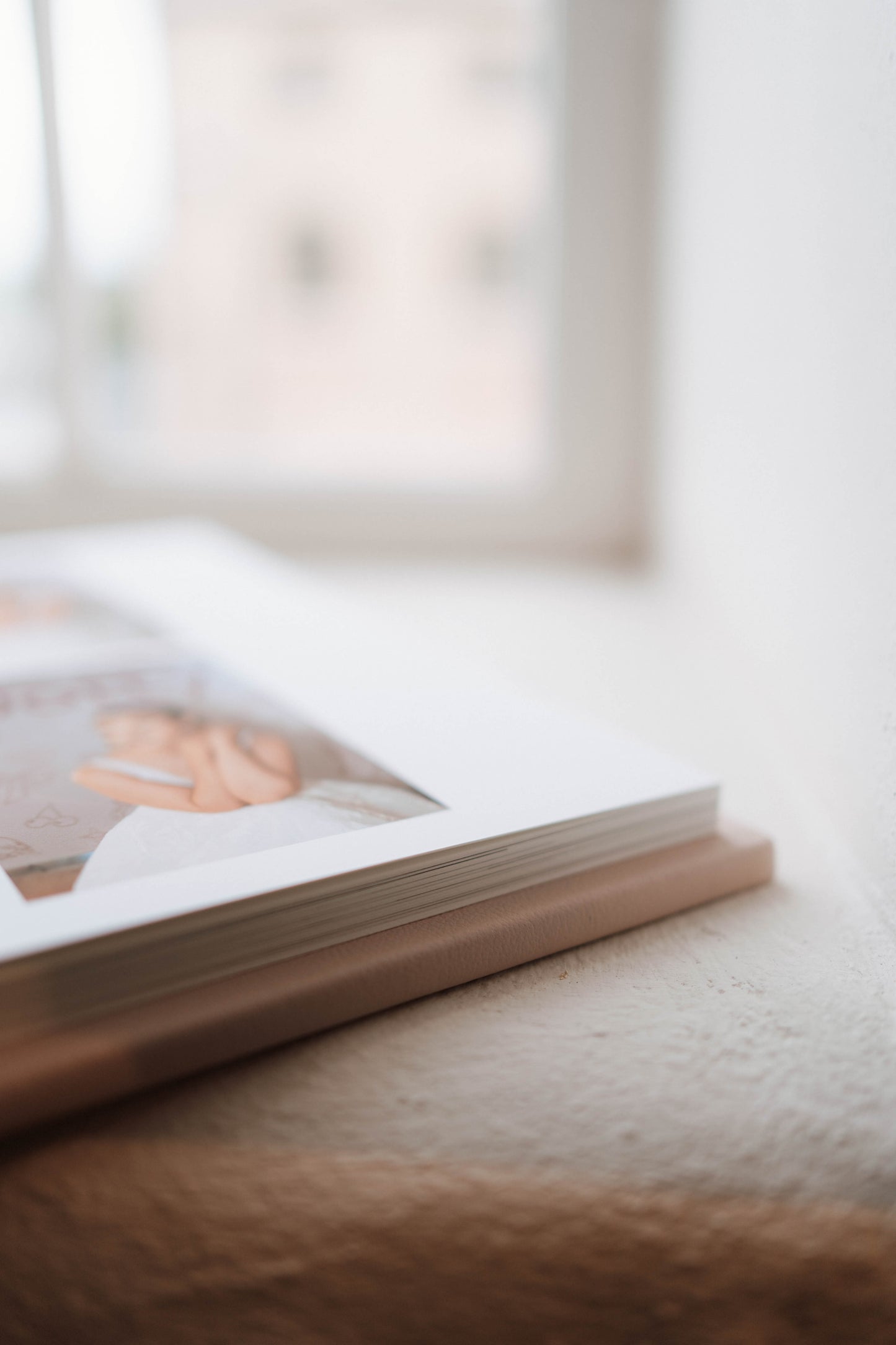 A close-up, shallow depth-of-field shot of a hardcover photo book lying on a textured beige surface near a window.