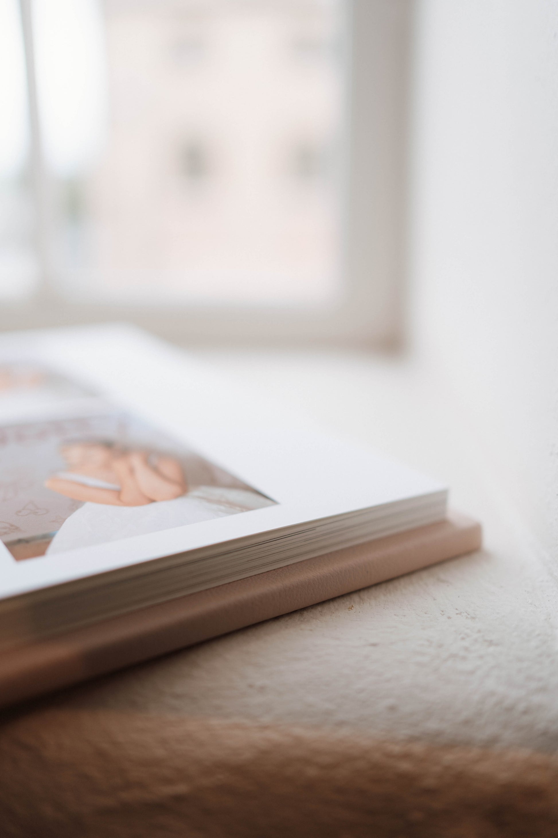 A close-up, shallow depth-of-field shot of a hardcover photo book lying on a textured beige surface near a window.