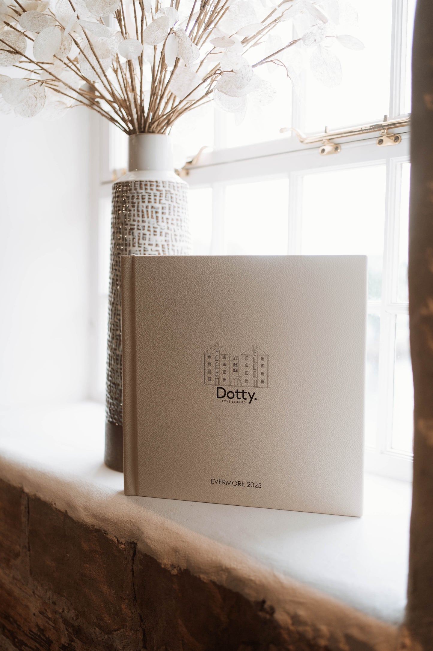 A cream-colored hardcover wedding album standing on a windowsill, with the brand name 'Dotty.' printed on the front.