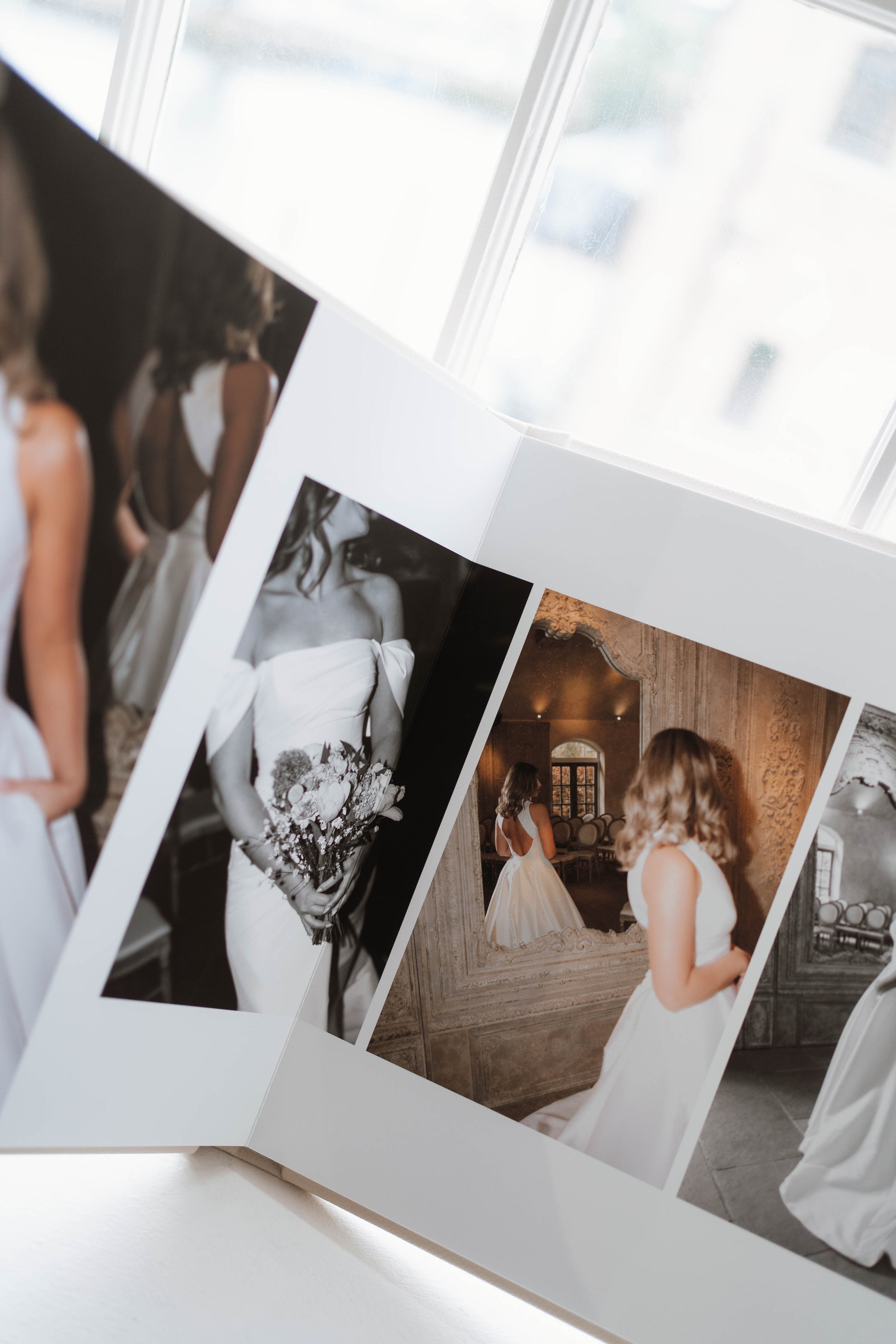 An open white photo album spread on a light surface, showing printed wedding photographs of brides in white dresses in a bright indoor setting.